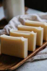 4 bars of naked goat milk soap on a wood dish with a dish towel underneath. a bottle of milk is in the background. 