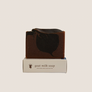 A bar of black amber and lavender goat milk soap on top of it's box with beige background