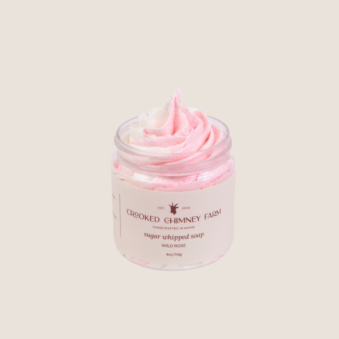 a 4oz jar of wild rose sugar whipped soap from crooked chimney farm. It is pink and white swirled, piped into a clear plastic jar with a tan and brown label. 