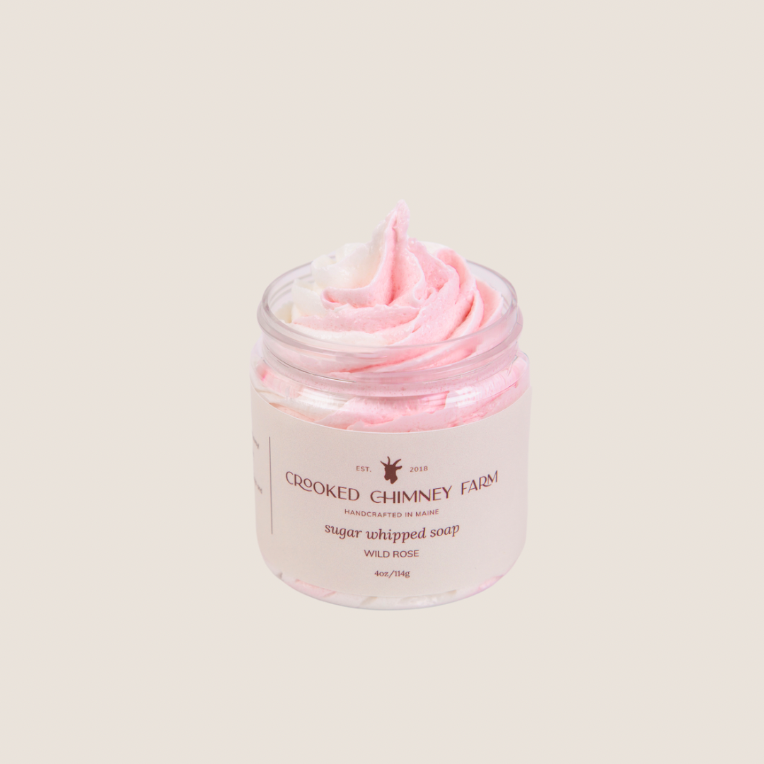 a 4oz jar of wild rose sugar whipped soap from crooked chimney farm. It is pink and white swirled, piped into a clear plastic jar with a tan and brown label.