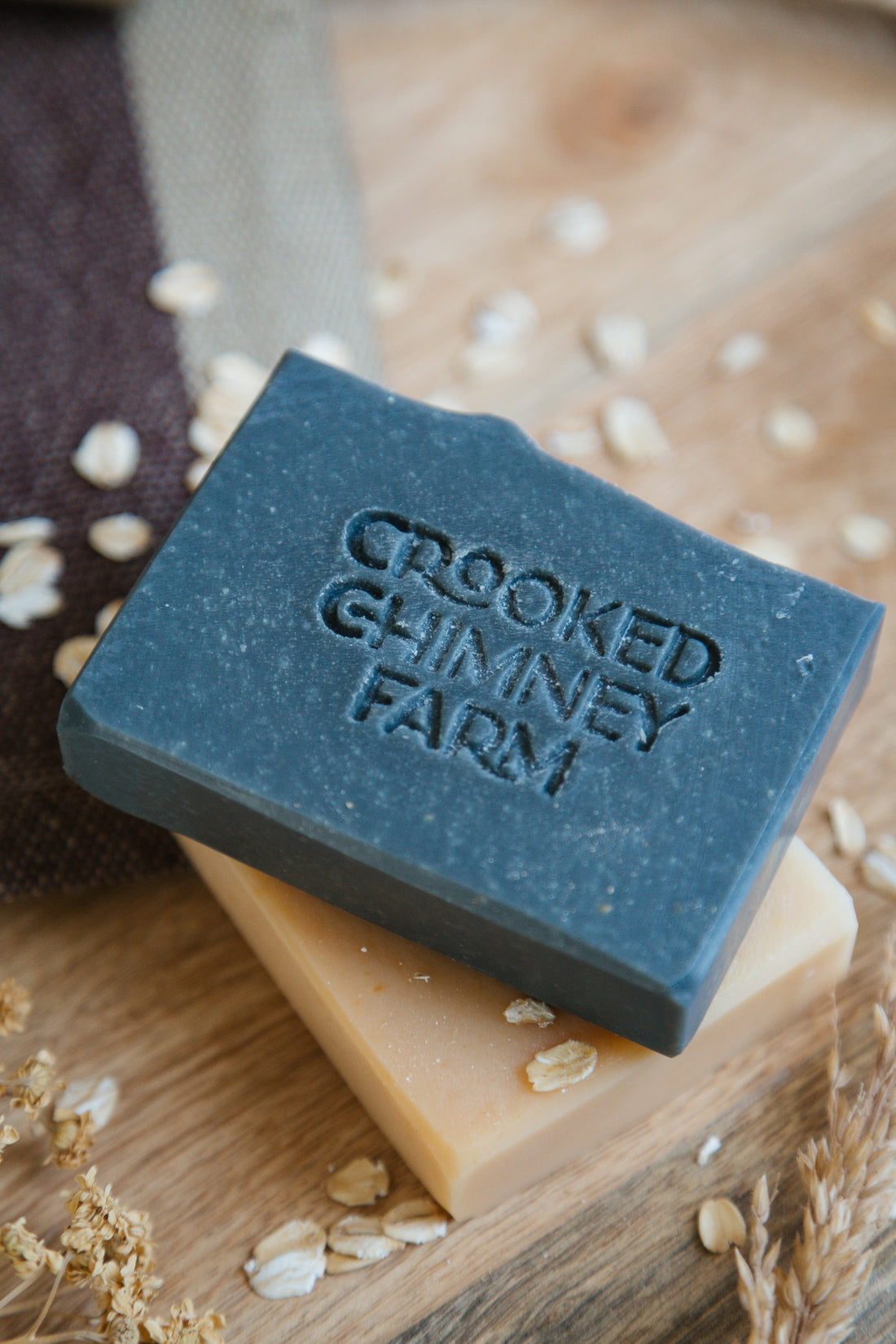 a bar of bramble soap on top of a bar of hearth soap on wood tray with dish towel and oats.