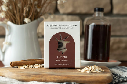 A box of Crooked Chimney Farm Hearth Handcrafted Goat Milk Soap stands on a wooden cutting board with oats and cinnamon scattered around it. A blurred bottle and dried flowers appear in the background against a warm brown wall.